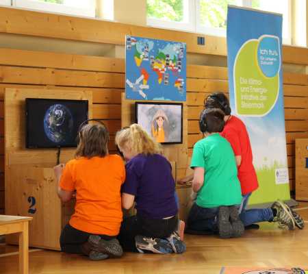 Kinder sitzen vor zwei Stationen mit Bildschirmen der Klimaversum-Wanderausstellung auf ihren Knien. Man sieht sie von hinten.